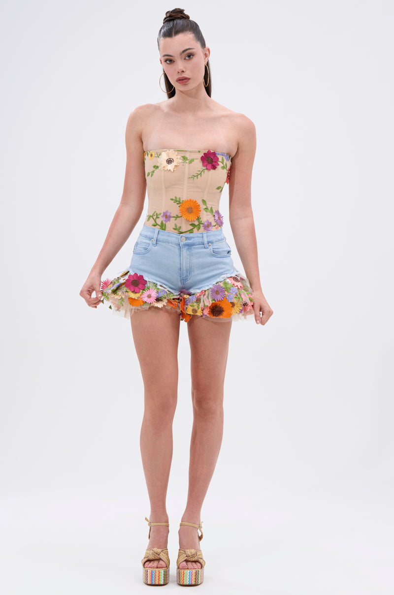 A woman with a high ponytail wears the VIVI FLORAL CORSET with sheer mesh overlay and colorful floral appliqués, paired with denim mini shorts and platform sandals, standing against a plain background.