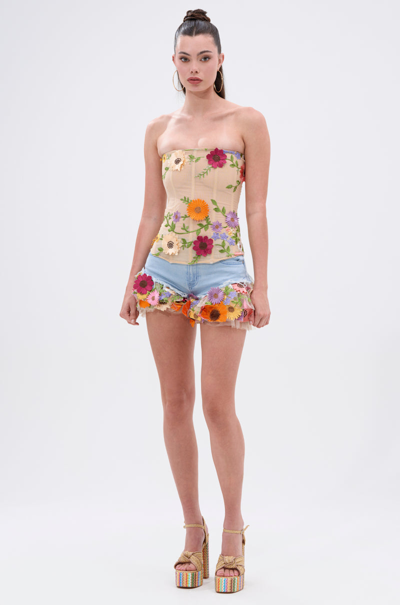 A woman with a high ponytail stands against a white background wearing the VIVI FLORAL CORSET, denim shorts with floral trim, rainbow platform heels, and gold hoop earrings.