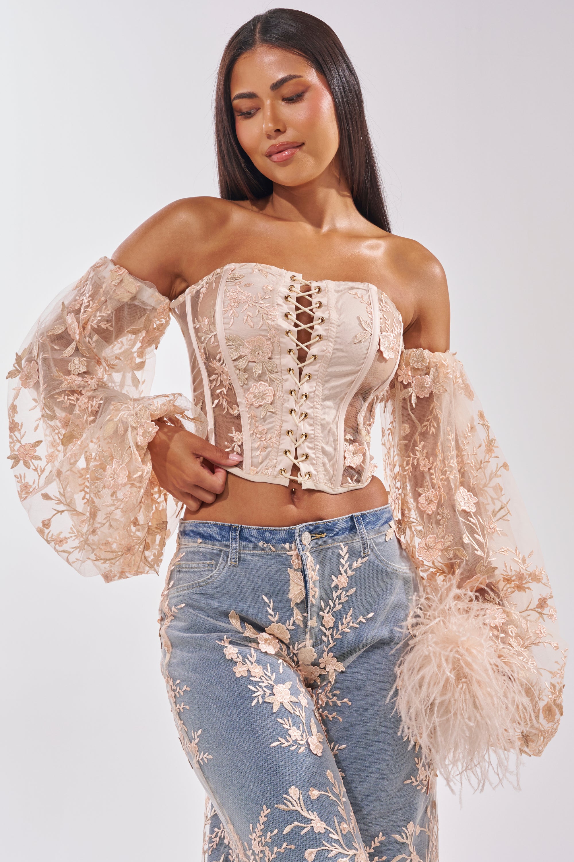 A woman poses against a plain white background in the FLY AWAY WITH ME FLORAL CORSET, featuring cream mesh, off-the-shoulder sleeves, and sheer floral embroidery, paired with embroidered blue jeans and a feathered accessory. 0