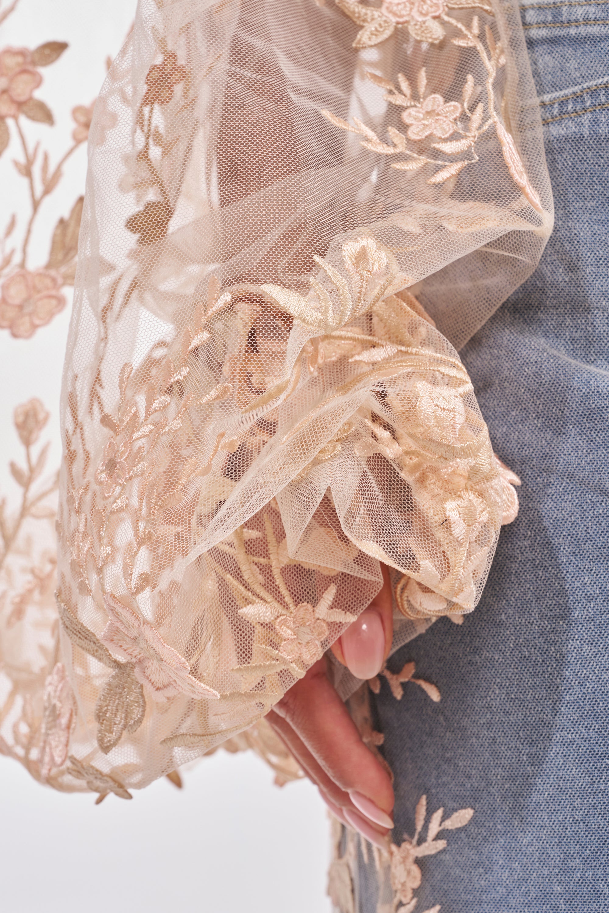 A close-up of a person wearing the FLY AWAY WITH ME FLORAL CORSET in soft beige mesh, detailed with delicate floral and leaf embroidery, with their hand resting near blue denim jeans. 10