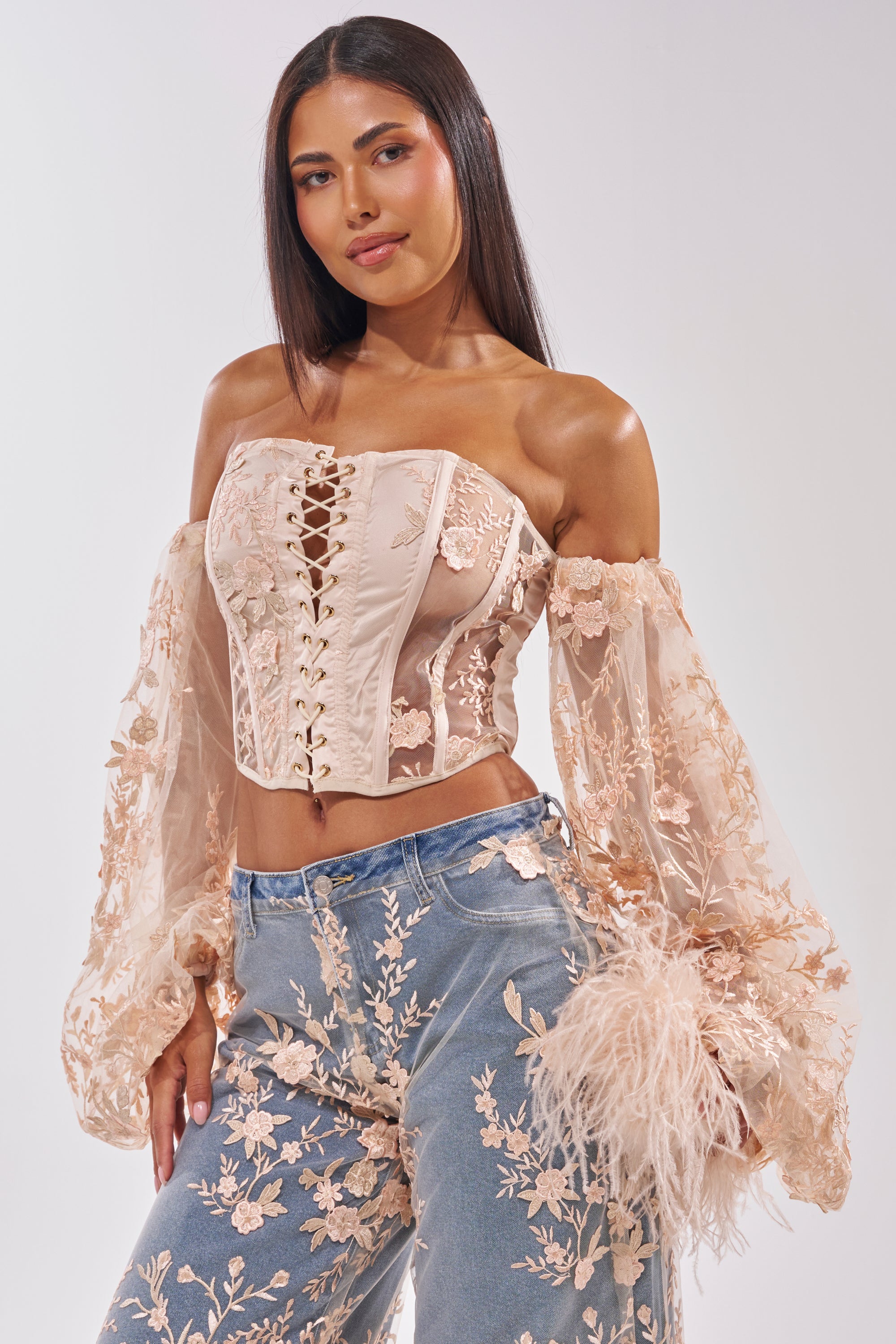 A woman with long, straight hair confidently models the FLY AWAY WITH ME FLORAL CORSET, a beige mesh off-shoulder top with sheer floral embroidery, paired with high-waisted blue jeans featuring matching floral accents. 2