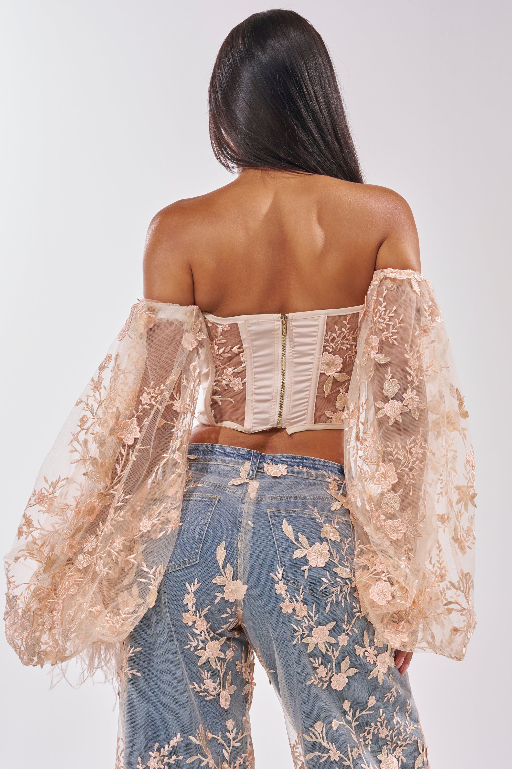 A woman faces away in the FLY AWAY WITH ME FLORAL CORSET, featuring sheer off-shoulder sleeves and puffy arms, styled with embroidered blue jeans. Her long dark hair falls down her back against a white backdrop. 4