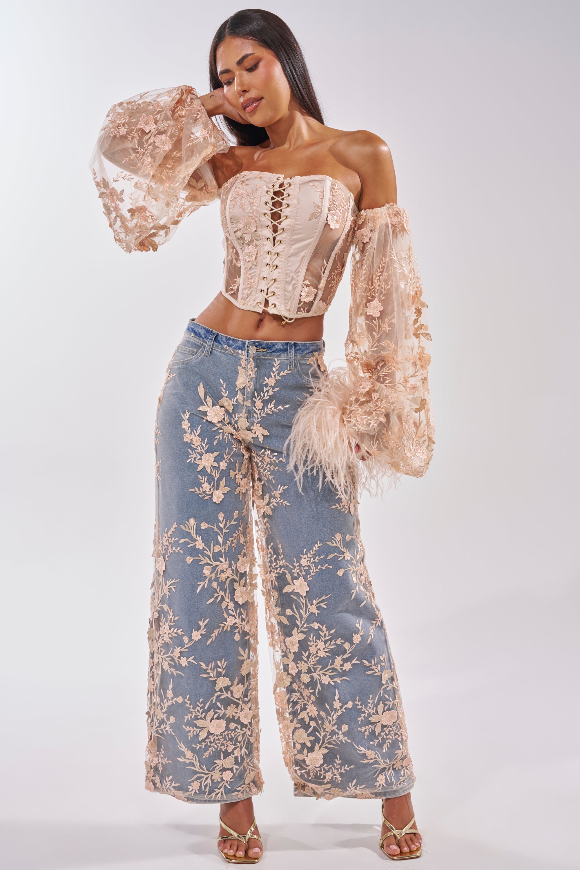 Woman poses in the FLY AWAY WITH ME FLORAL CORSET, a beige mesh top with off-the-shoulder sleeves and sheer accents, styled with wide-leg blue jeans featuring beige floral embroidery and high-heeled sandals. 5