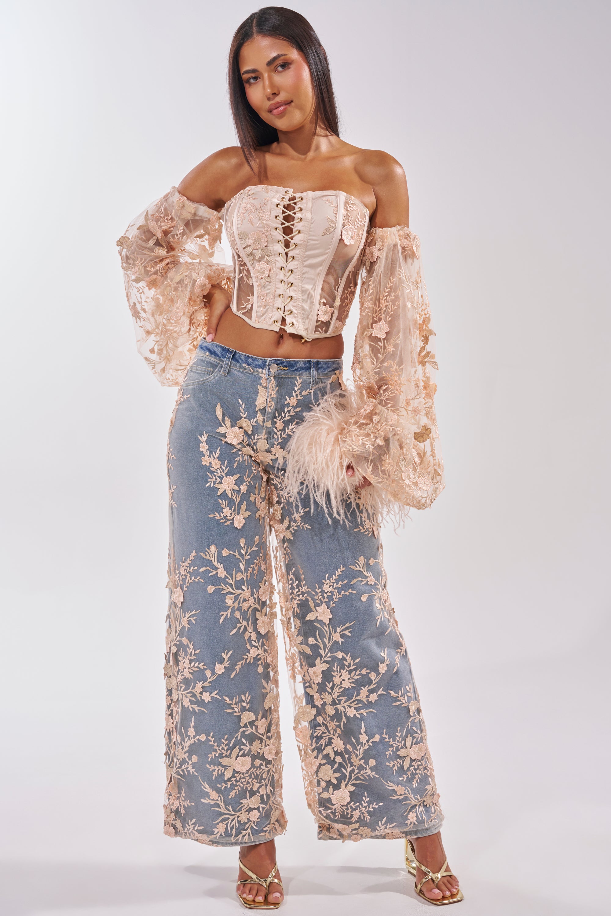 A woman wears the FLY AWAY WITH ME FLORAL CORSET with off-the-shoulder sheer sleeves and wide-leg denim jeans featuring matching floral embroidery, paired with gold heels. She stands against a plain, light-colored background. 7