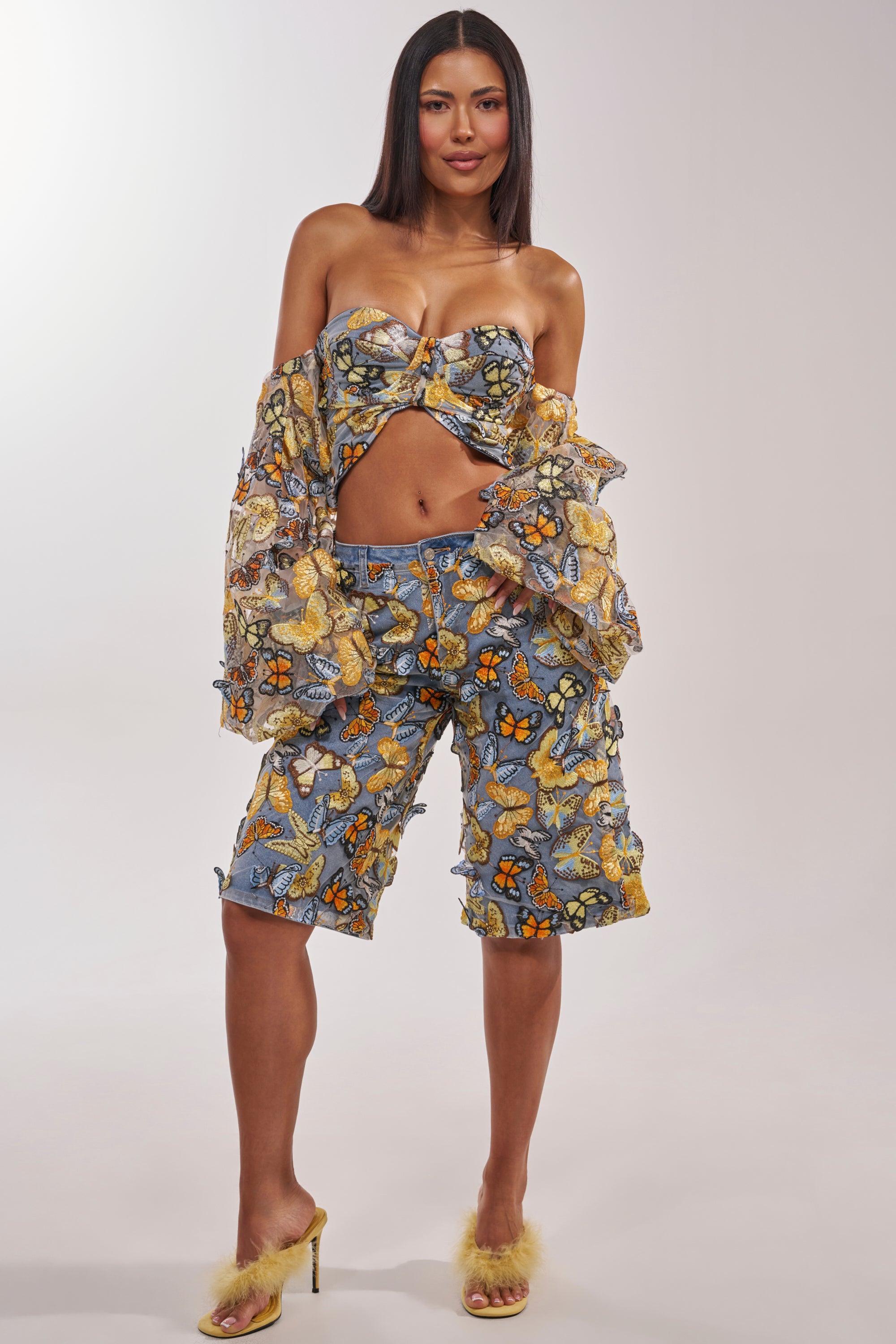 A woman exudes confidence in the BUTTERFLY EFFECT BLOUSE—a strapless corset style—paired with matching shorts and a sheer jacket. She finishes the look with yellow feathered heels, standing against a plain white background. 9