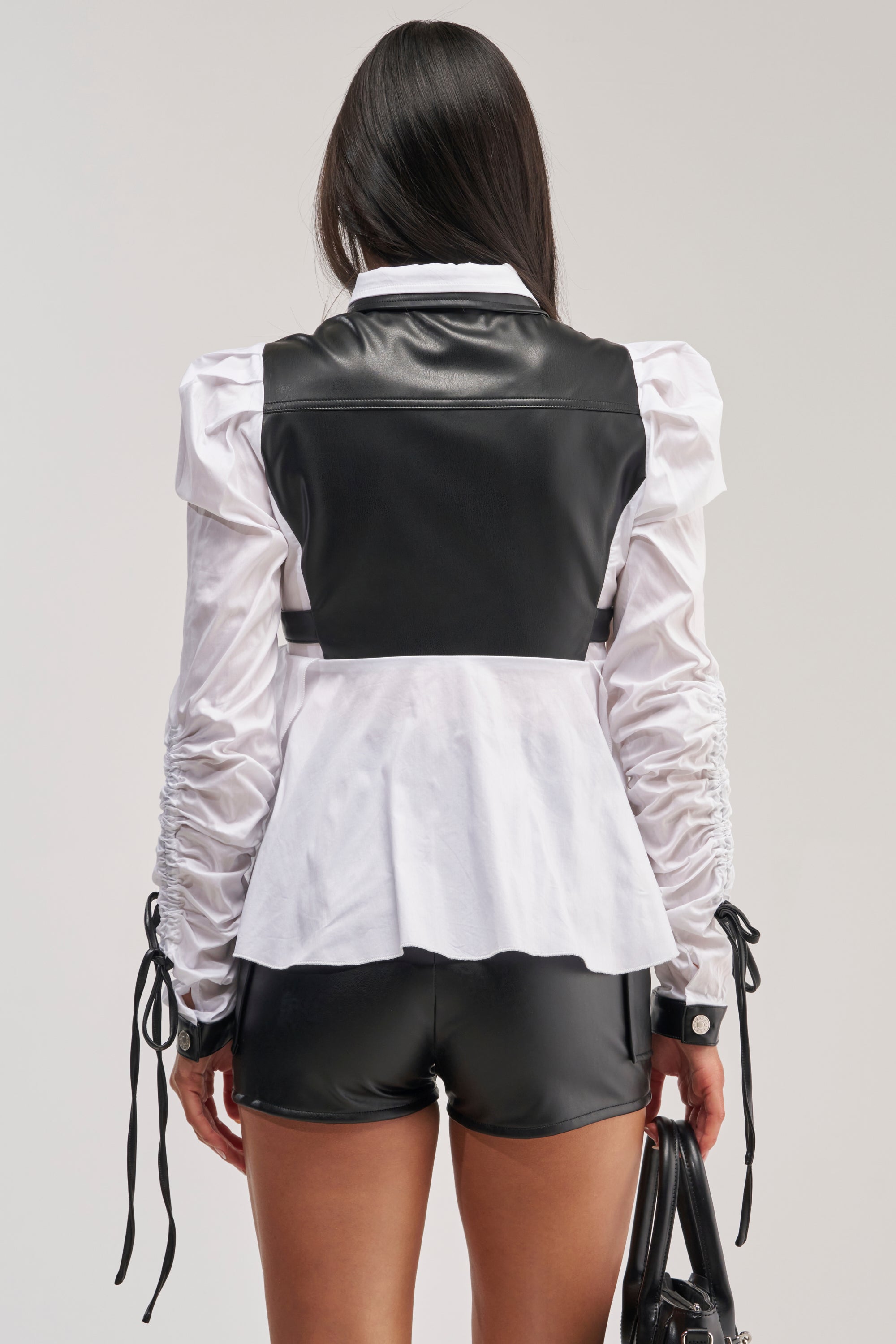 A woman stands facing away, wearing the ALWAYS FABULOUS FAUX LEATHER DETAIL BUTTON DOWN BLOUSE—a white long-sleeve top with puffed sleeves and black faux leather detailing—paired with matching shorts, and holding a black handbag.