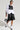 A fashionable person stands confidently wearing a layered outfit. They have the ALWAYS FABULOUS Faux Leather Detail Button Down Blouse, adorned with ruched drawstring sleeves and featuring a black faux leather bodice over it. Complementing their look are black leather shorts, black pointed heels paired with white socks, and they hold a chic white and black handbag. Their ensemble is completed with stylish glasses and hoop earrings.