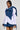 A person wearing a stylish ensemble featuring the MELODY DENIM TIE POPLIN BLOUSE, which includes a white poplin blouse accented with an oversized denim bow, paired with blue shorts. The plain background emphasizes the individual's confident expression.