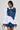 A woman confidently poses against a plain background, wearing the MELODY DENIM TIE POPLIN BLOUSE. The blouse features an oversized denim bow on her left shoulder, and she complements her stylish outfit with short black hair.