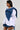 A person stands against a plain background wearing a stylized outfit that includes the MELODY DENIM TIE POPLIN BLOUSE with long sleeves, an oversized denim bow, and denim bottoms. They have a short, sleek hairstyle and are looking to the left.