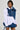 A person stylishly dressed in the MELODY DENIM TIE POPLIN BLOUSE, showcasing wide ruffled sleeves and an oversized bow on one shoulder, complemented by a blue denim vest and matching denim shorts against a simple, neutral background.