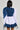 A person with shoulder-length black hair is seen from behind wearing a layered outfit: the MELODY DENIM TIE POPLIN BLOUSE featuring ruched sleeves, paired with a denim vest and skirt. They're holding a denim jacket draped over one shoulder, and an oversized bow adds an elegant touch.