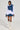The MELODY DENIM TIE POPLIN BLOUSE, worn over a chic denim and white layered dress with an oversized denim bow on one shoulder, perfectly matches stylish white lace-up knee-high boots. The outfit's fashion-forward design, complete with a white collar and long sleeves, takes center stage against the plain background.