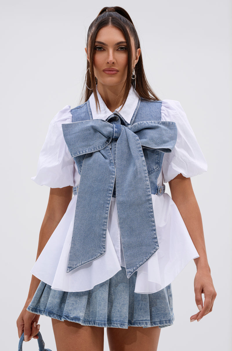 A woman wears the FAITH SHORT SLEEVE DENIM TIE POPLIN BLOUSE TOP featuring a large denim bow, paired with a pleated denim mini skirt and small denim bag. Her long brown hair is in a high ponytail against a plain light background. 0