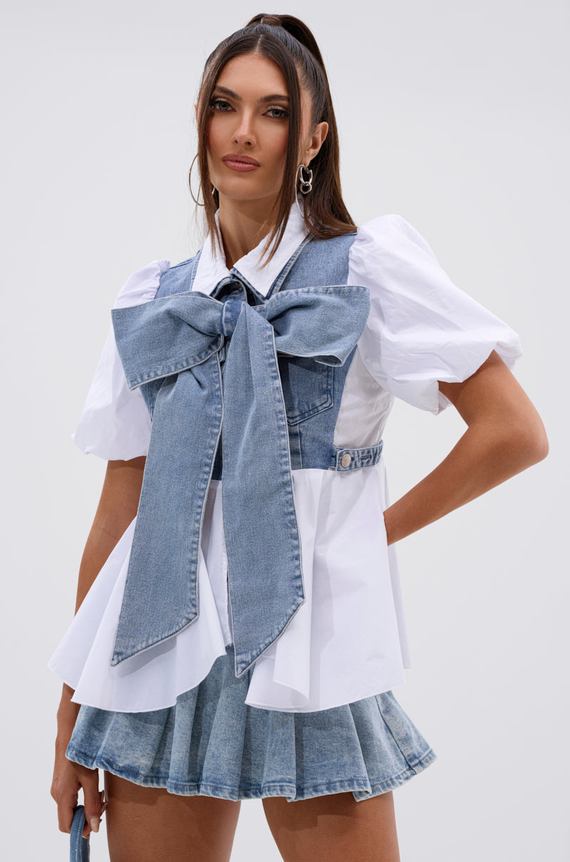 A woman stands against a plain light background, wearing the FAITH SHORT SLEEVE DENIM TIE POPLIN BLOUSE TOP styled with a large denim bow and corset, paired with a pleated light blue denim mini skirt. 1