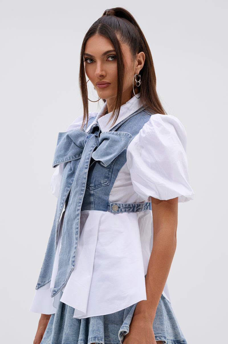 A woman with long brown hair in a high ponytail wears the FAITH SHORT SLEEVE DENIM TIE POPLIN BLOUSE TOP with a large denim bow, layered vest, matching denim skirt, hoop earrings, and has a neutral expression. 3