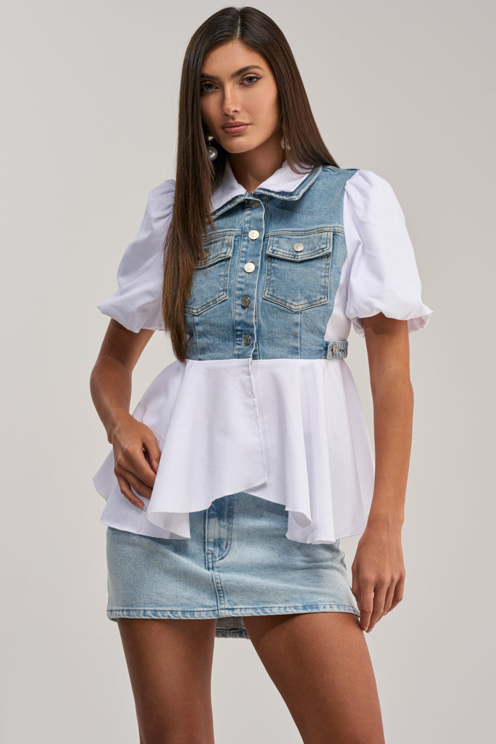 A woman wears the HOPELESS ROMANTIC DENIM BLOUSE, a stylish denim vest layered over a poplin peplum top with puffed sleeves, paired with a light blue denim mini skirt against a plain background.