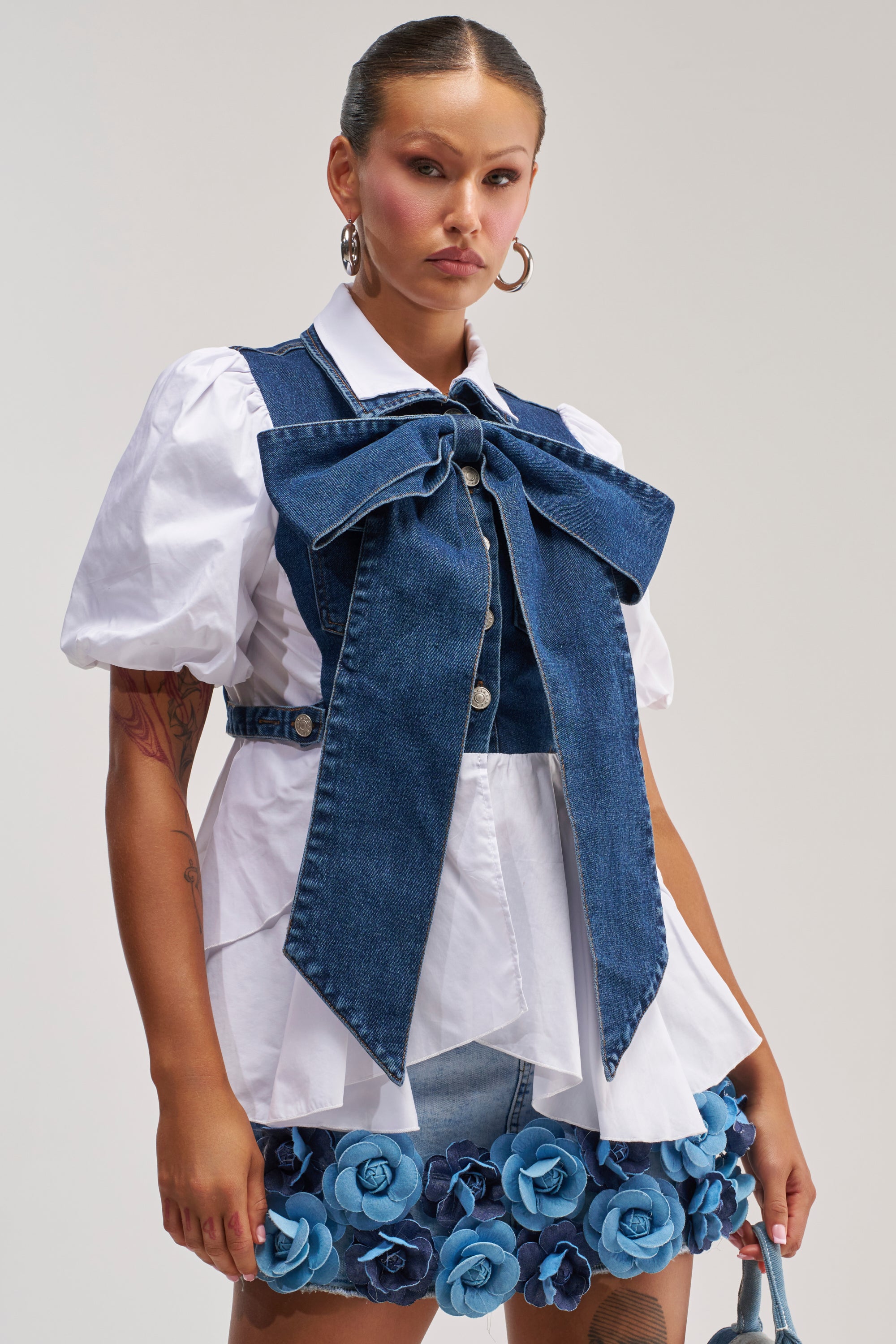 Dressed in the FAITH SHORT SLEEVE DARK BLUE DENIM TIE POPLIN BLOUSE, this woman creates a statement summer look by pairing it with a denim mini skirt adorned with blue 3D flowers, hoop earrings, and a denim handbag—her outfit stands out against the neutral background.