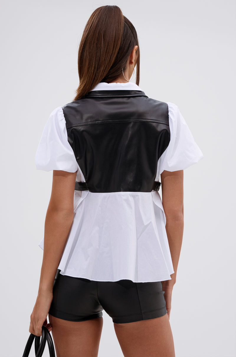 A woman stands with her back to the camera in a plain light gray setting, wearing the NEW DRESS CODE LEATHER AND POPLIN BUTTON DOWN SHIRT over black shorts, holding a handbag in her left hand, her straight hair pulled back. 4