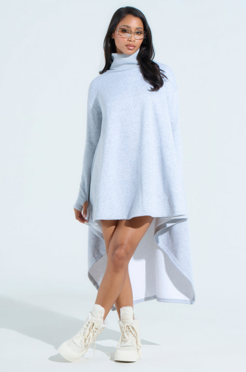 A person in the LAINIE Turtleneck High Low Sweatshirt Dress (light gray, fleece-lined) stands confidently against a plain background, wearing cream-colored boots and glasses with a relaxed posture, one leg slightly forward.