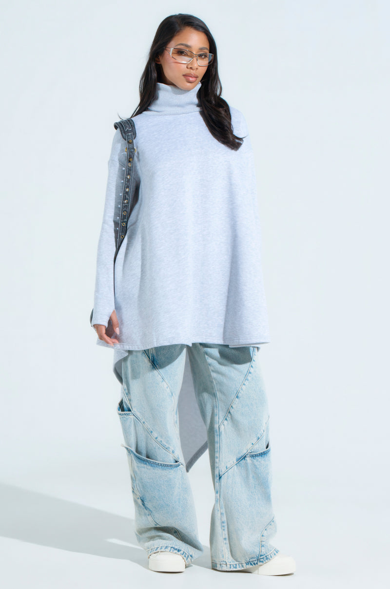Dressed in cozy chic fashion, a person wears a Lainie Turtleneck High Low Sweatshirt Dress in light gray, oversized light blue jeans with asymmetrical seams, and white sneakers. They accessorize with sunglasses and a black shoulder bag against a plain light background.