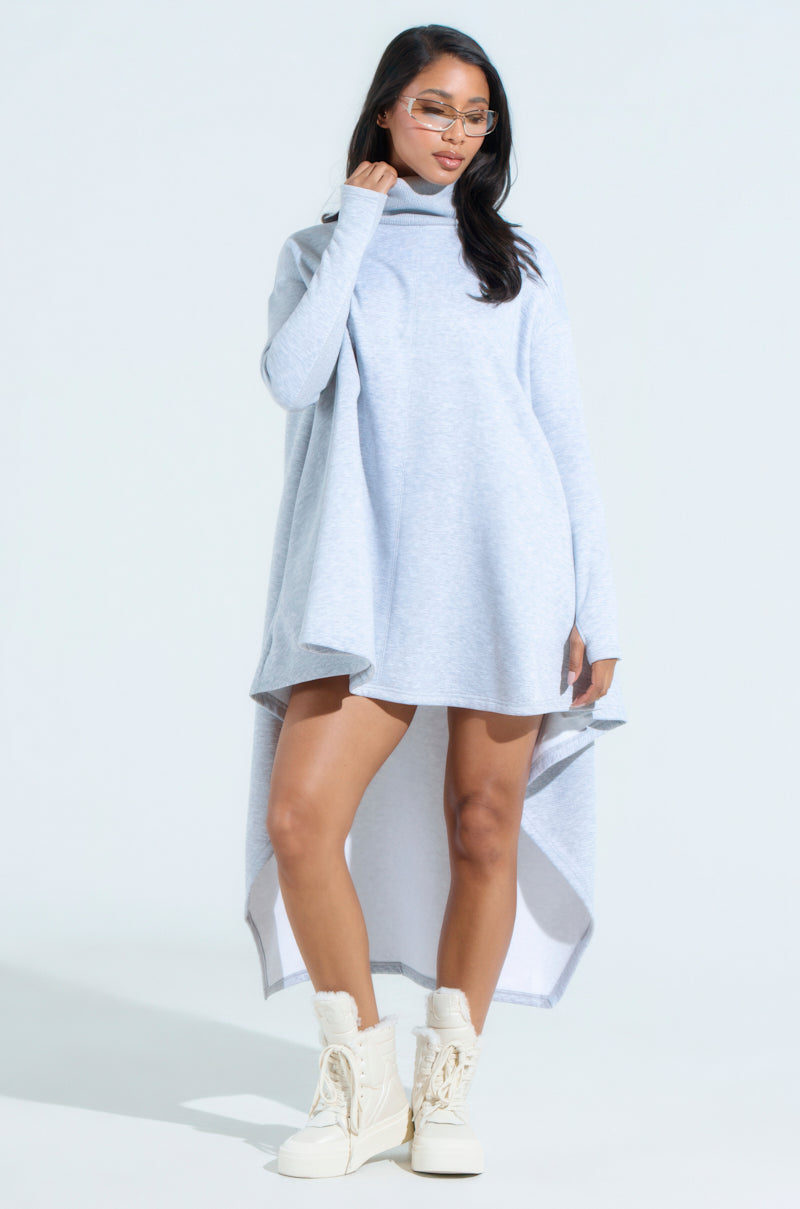 A person poses against a plain background wearing the LAINIE TURTLENECK HIGH LOW SWEATSHIRT DRESS. They complete the cozy chic look with long hair, glasses, and white fur-trimmed boots, showcasing the dress's asymmetrical hemline.