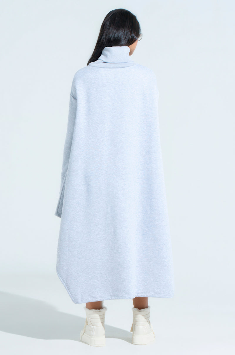 A person with long dark hair stands facing away, wearing the LAINIE TURTLENECK HIGH LOW SWEATSHIRT DRESS in light gray and white ankle boots, embodying cozy chic fashion against a plain backdrop.
