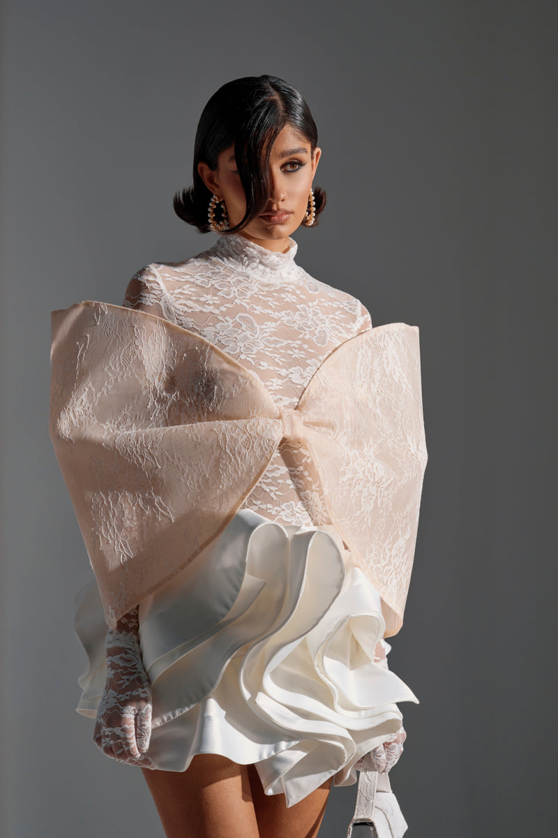 A woman in the white I'M A STAR LACE BOW TOP with sheer sleeves and a large pale pink bow wears a ruffled mini skirt, has short styled dark hair, statement earrings, and carries a white handbag.