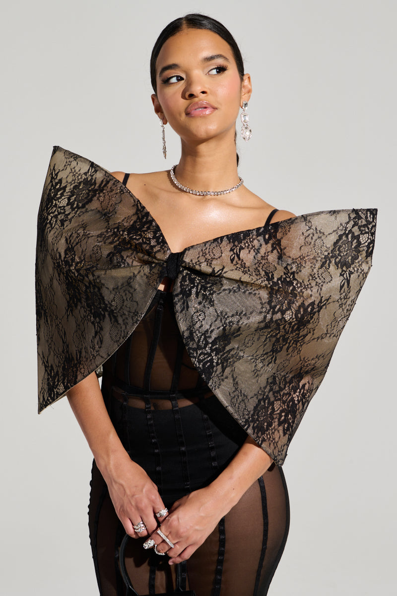 A woman poses in a chic ensemble featuring the ALL THE ATTENTION DRAMATIC BOW TOP, crafted with a sheer black lace fabric and an eye-catching bow across the shoulders. Her hair is elegantly pulled back, highlighting her earrings, choker, and multiple rings. The outfit captivates with its alluring open back, set against a simple light-colored background.
