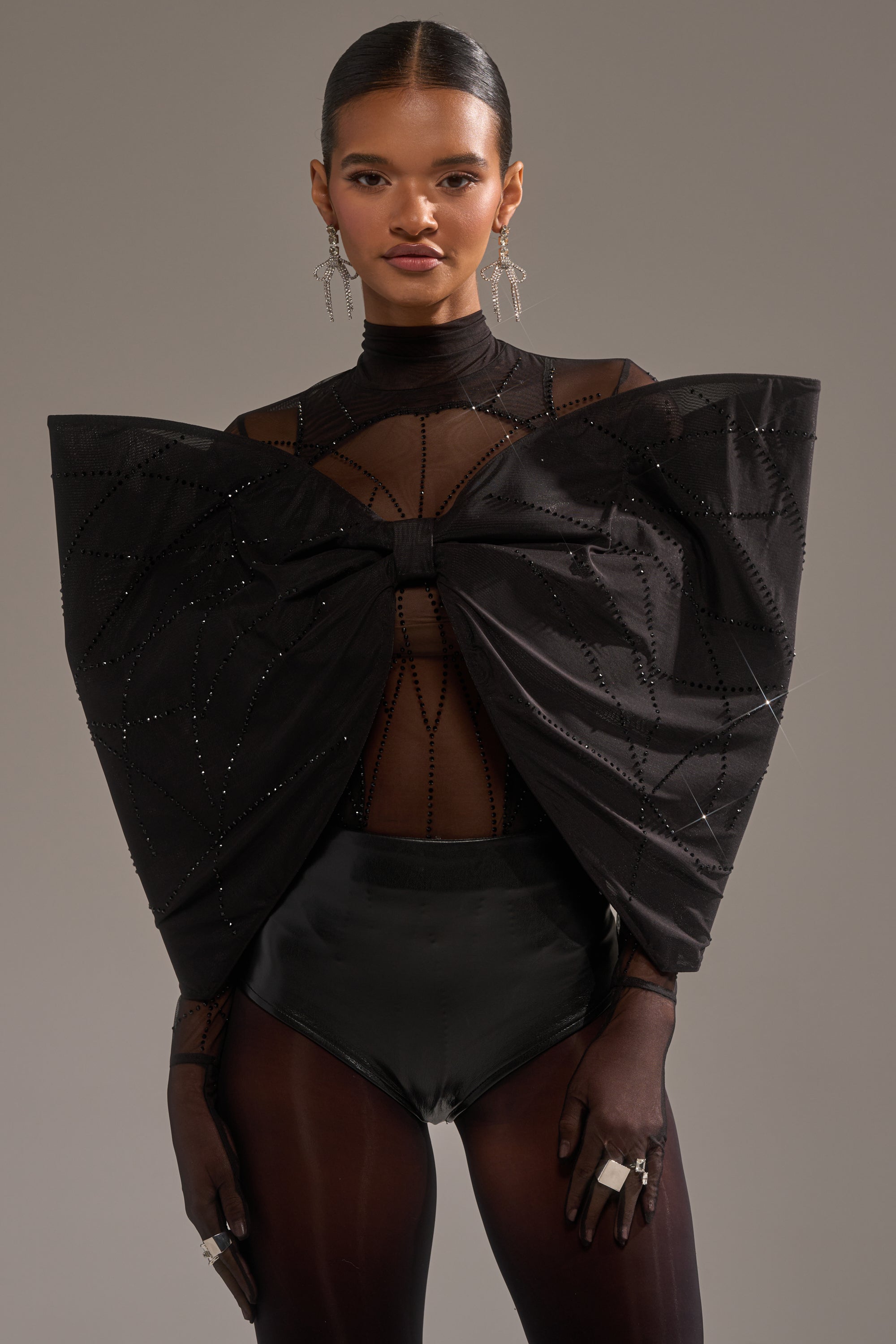 A woman wears the SPIDER GIRL BOW TOP, a sheer black top with a sculptural studded bow, styled with black high-waisted bottoms, sheer gloves, bold rings, and long silver earrings against a neutral background.