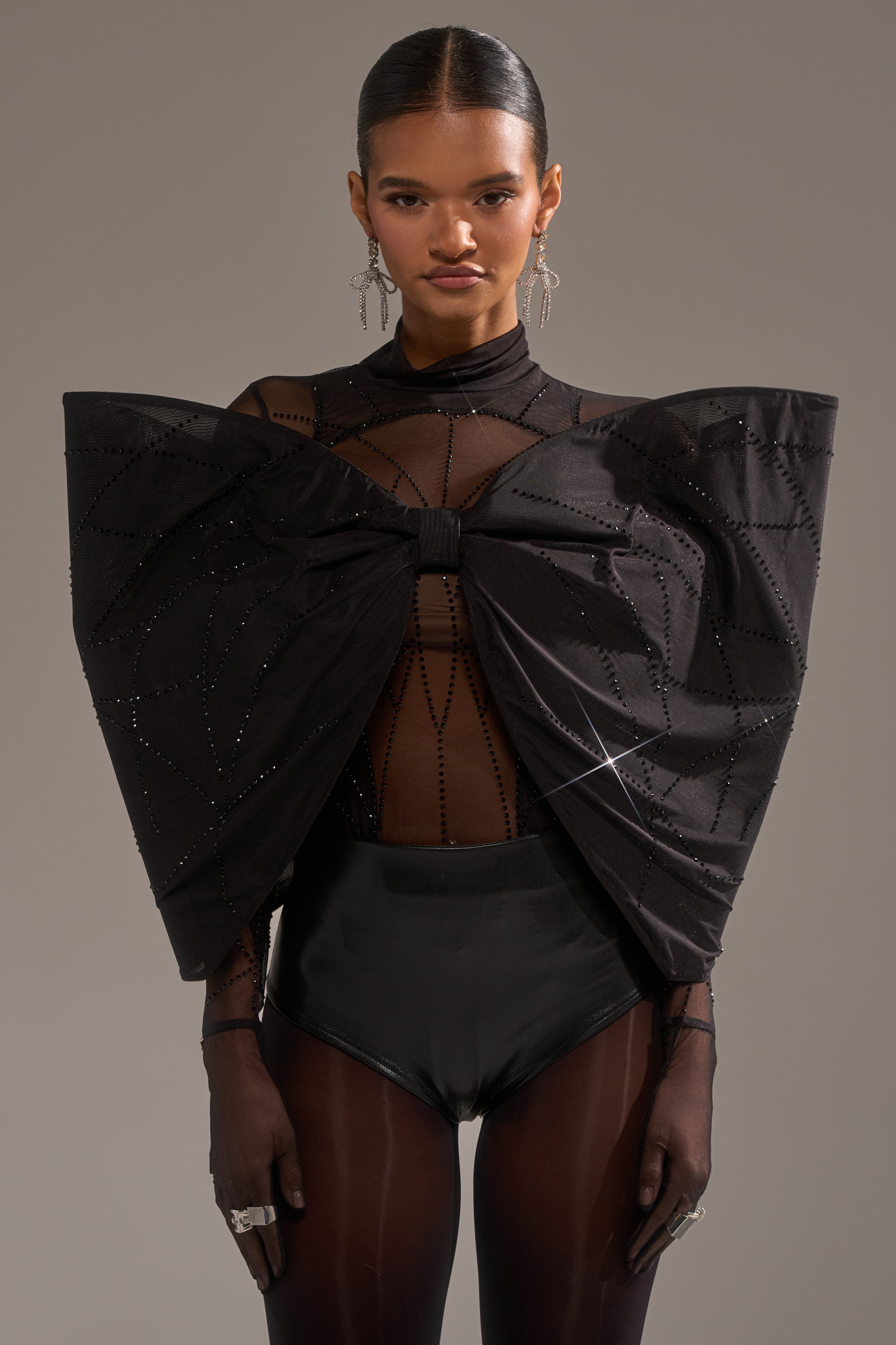 A woman in the SPIDER GIRL BOW TOP with sculptural geometric patterns, black high-waisted bottoms, sheer gloves, statement earrings, and rings stands against a plain background.