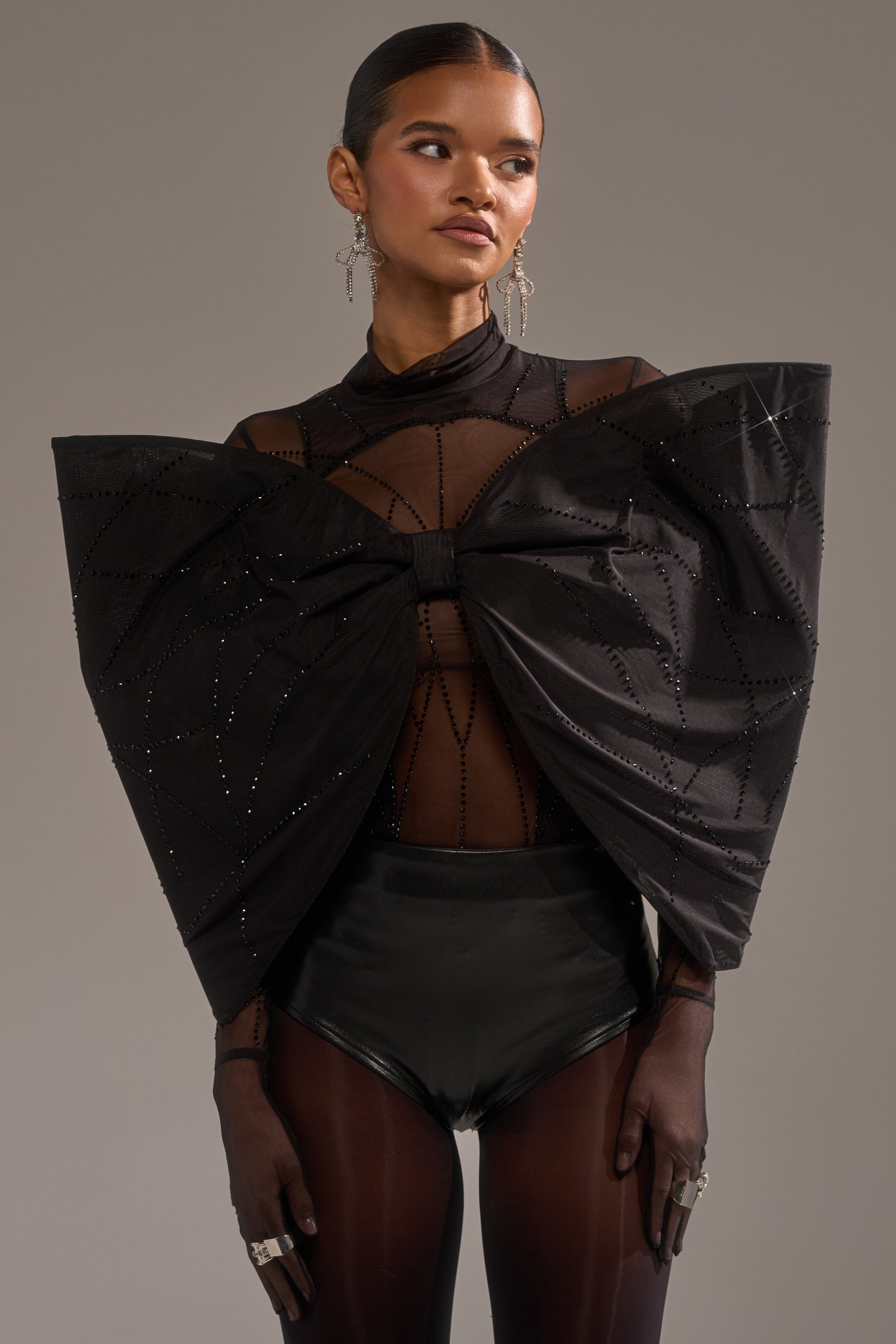 A woman wears the SPIDER GIRL BOW TOP—a sheer black top with a large, beaded sculptural bow—styled with high-waisted shorts, sheer gloves, statement rings, and dangling earrings against a neutral background.
