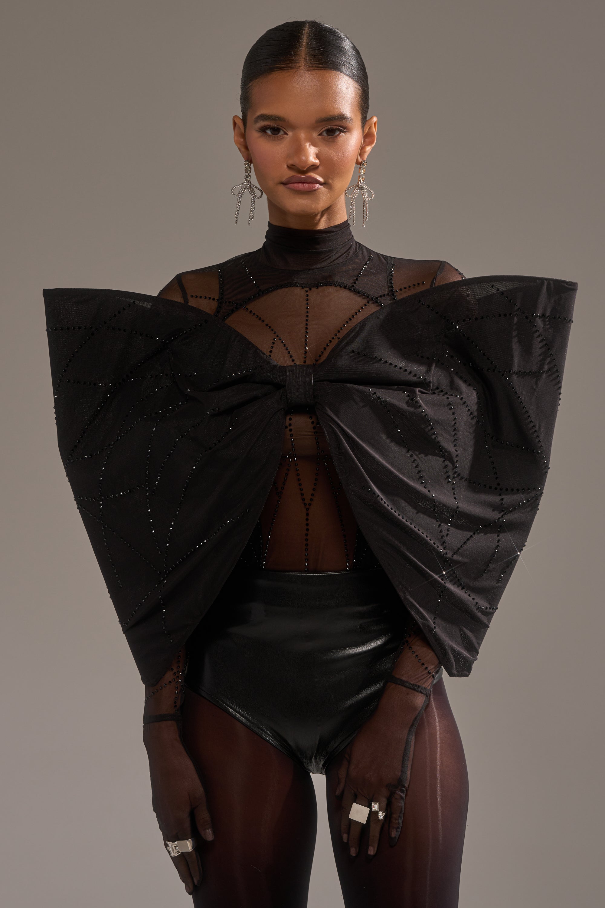 A woman poses confidently in the SPIDER GIRL BOW TOP, a sculptural black bow adorned with rhinestones, layered over a sheer black bodysuit. She pairs the look with sleek hair, statement earrings, and rings against a plain background.