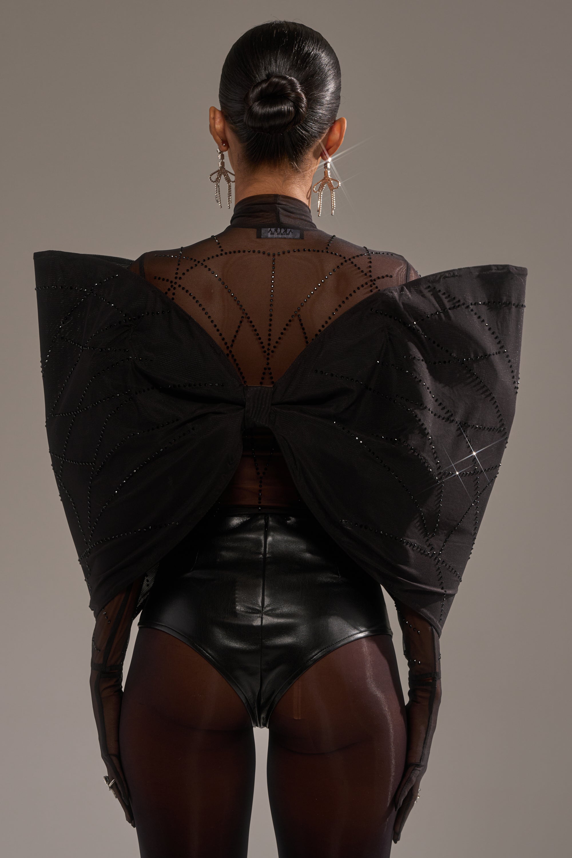 A person stands facing away, wearing the SPIDER GIRL BOW TOP—sheer black with rhinestone embellishments and a large sculptural bow—paired with black high-waisted shorts, sheer tights, gloves, bold earrings, and a sleek bun.