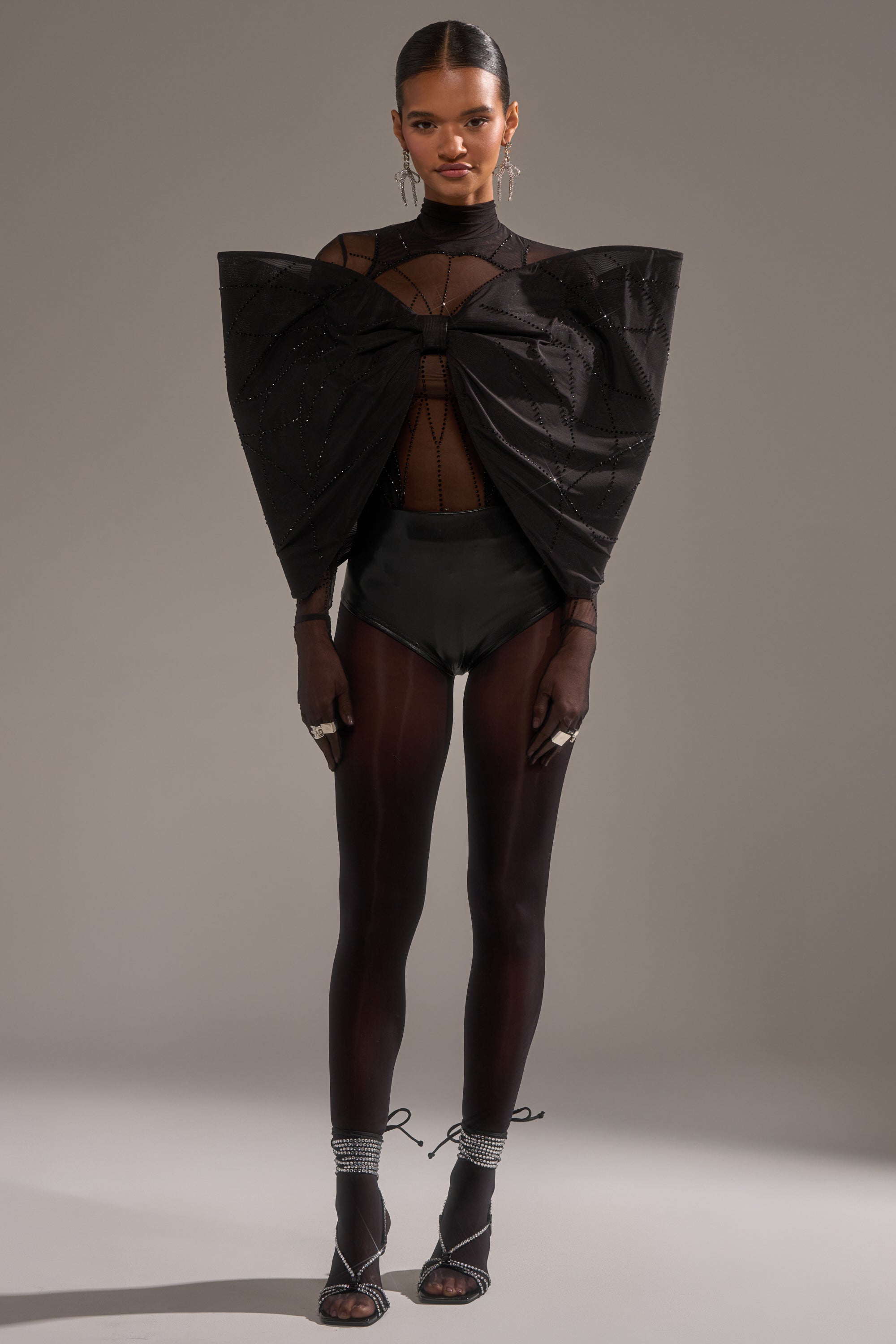 A woman poses confidently in the SPIDER GIRL BOW TOP, featuring dramatic sheer bow sleeves, paired with high-waisted bottoms, sheer tights, strappy heels, and statement jewelry against a simple backdrop.