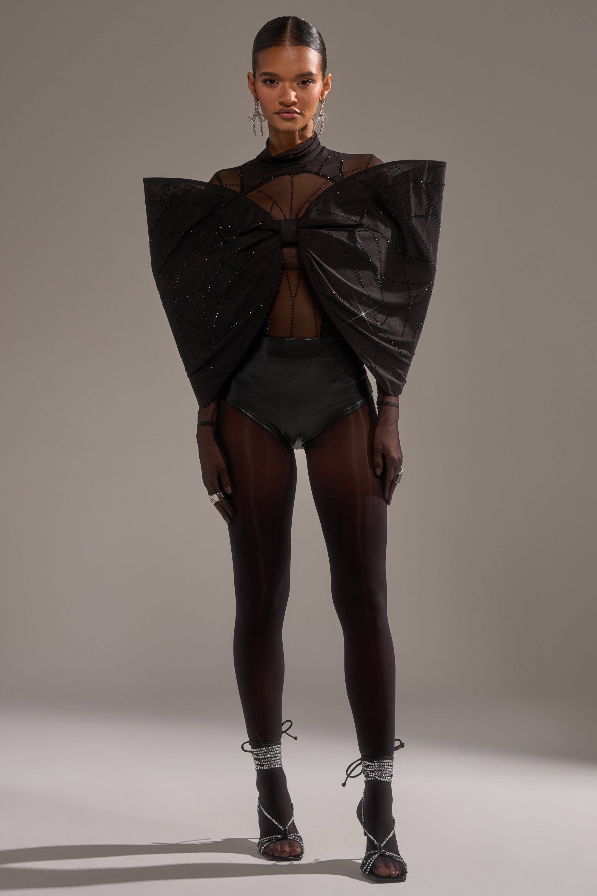 A woman faces forward in the SPIDER GIRL BOW TOP featuring a sculptural chest bow, paired with sheer black tights, high-waisted briefs, strappy heels, slicked-back hair, and statement earrings.