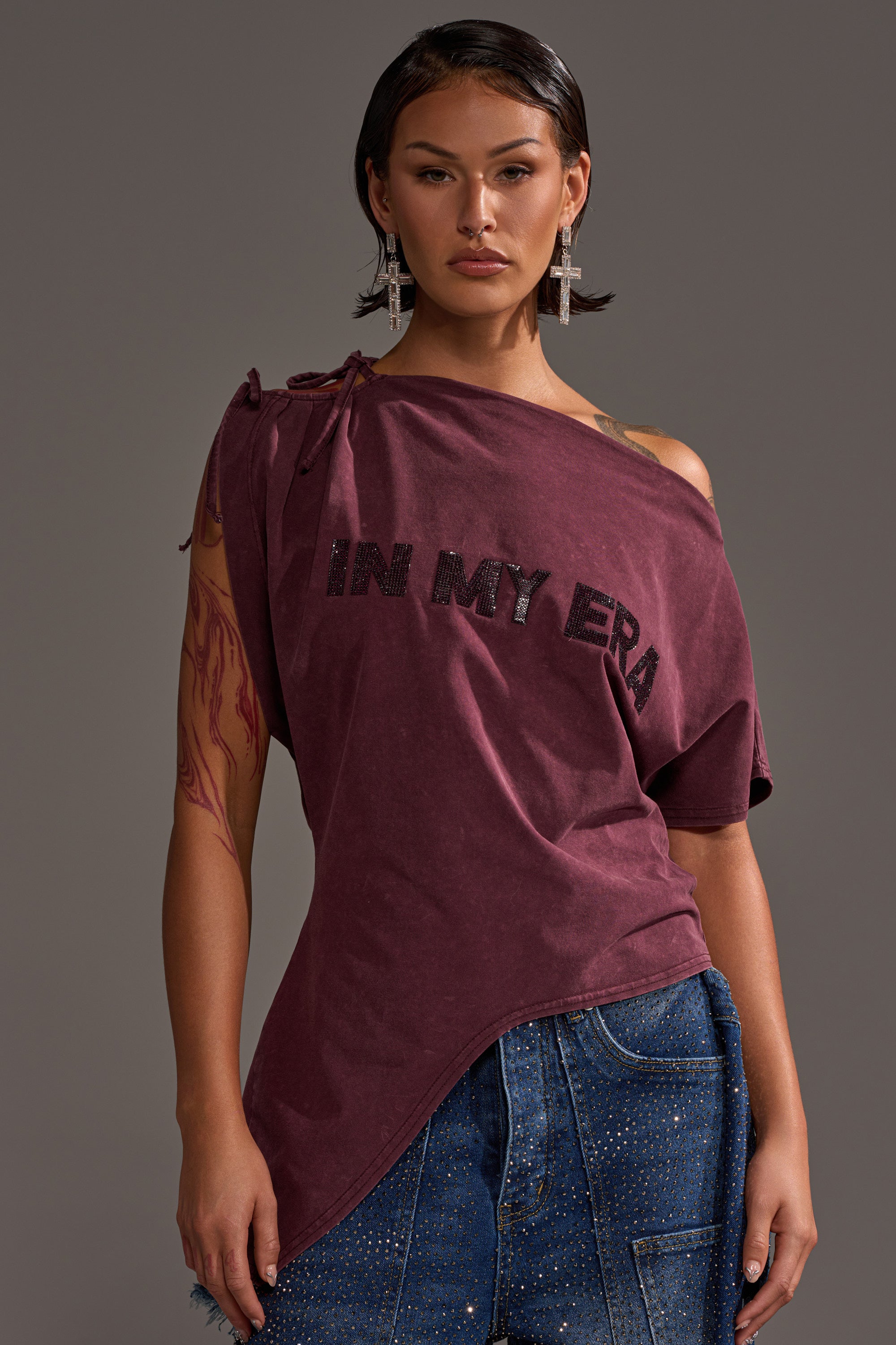 A woman stands against a plain background in the maroon IN MY ERA OFF-THE-SHOULDER TEE with black lettering, paired with cross-shaped embellished earrings and blue denim jeans with rhinestone detailing.