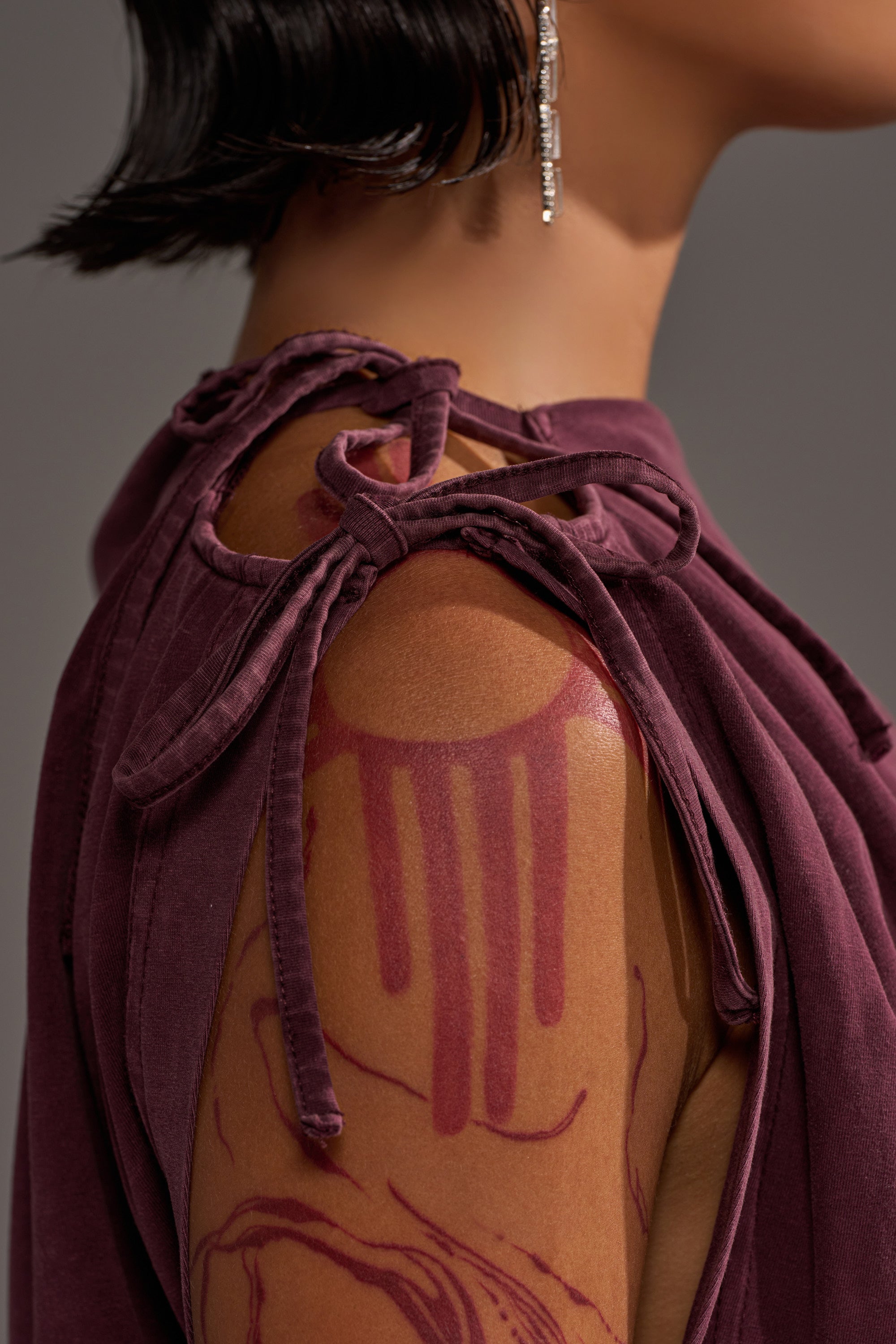 A person with short, dark hair and a dangling earring wears the IN MY ERA OFF-THE-SHOULDER TEE in deep purple, featuring fabric shoulder ties and revealing a maroon abstract tattoo on their upper arm.
