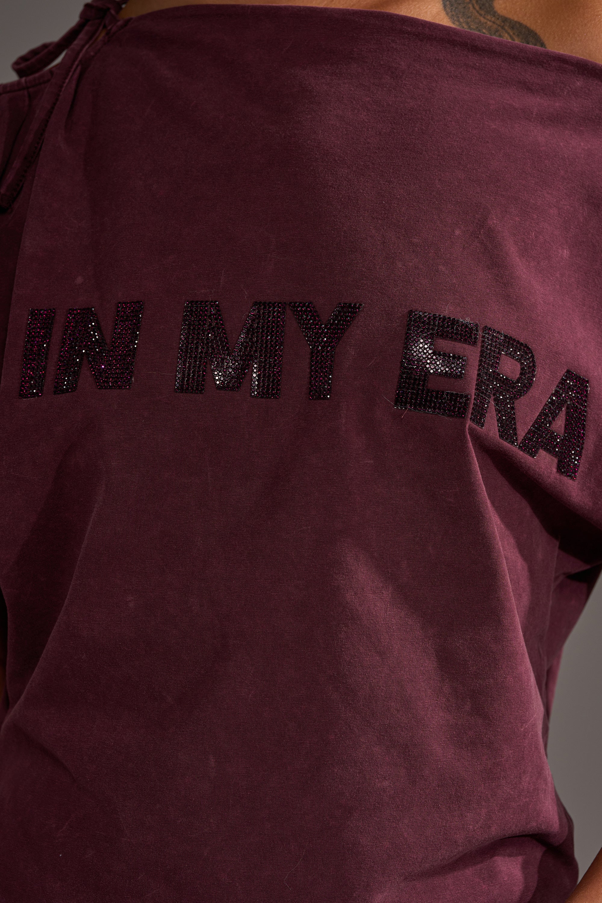 A close-up of the maroon IN MY ERA OFF-THE-SHOULDER TEE featuring black sequined "IN MY ERA" lettering across the back.