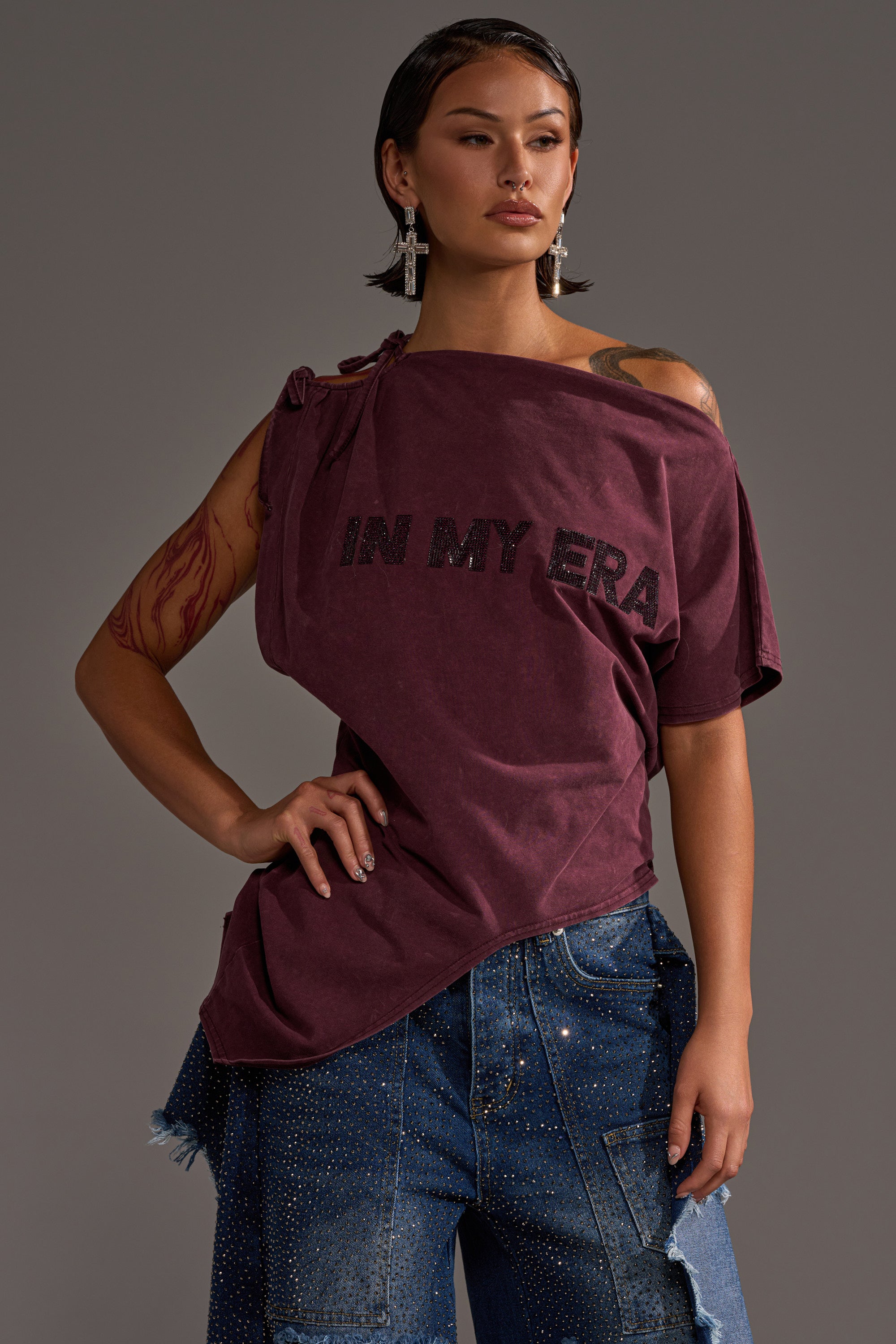 A woman poses confidently in the IN MY ERA OFF-THE-SHOULDER TEE, maroon with bold black lettering, paired with rhinestone-detailed blue denim pants, silver earrings, and showcasing a visible arm tattoo.