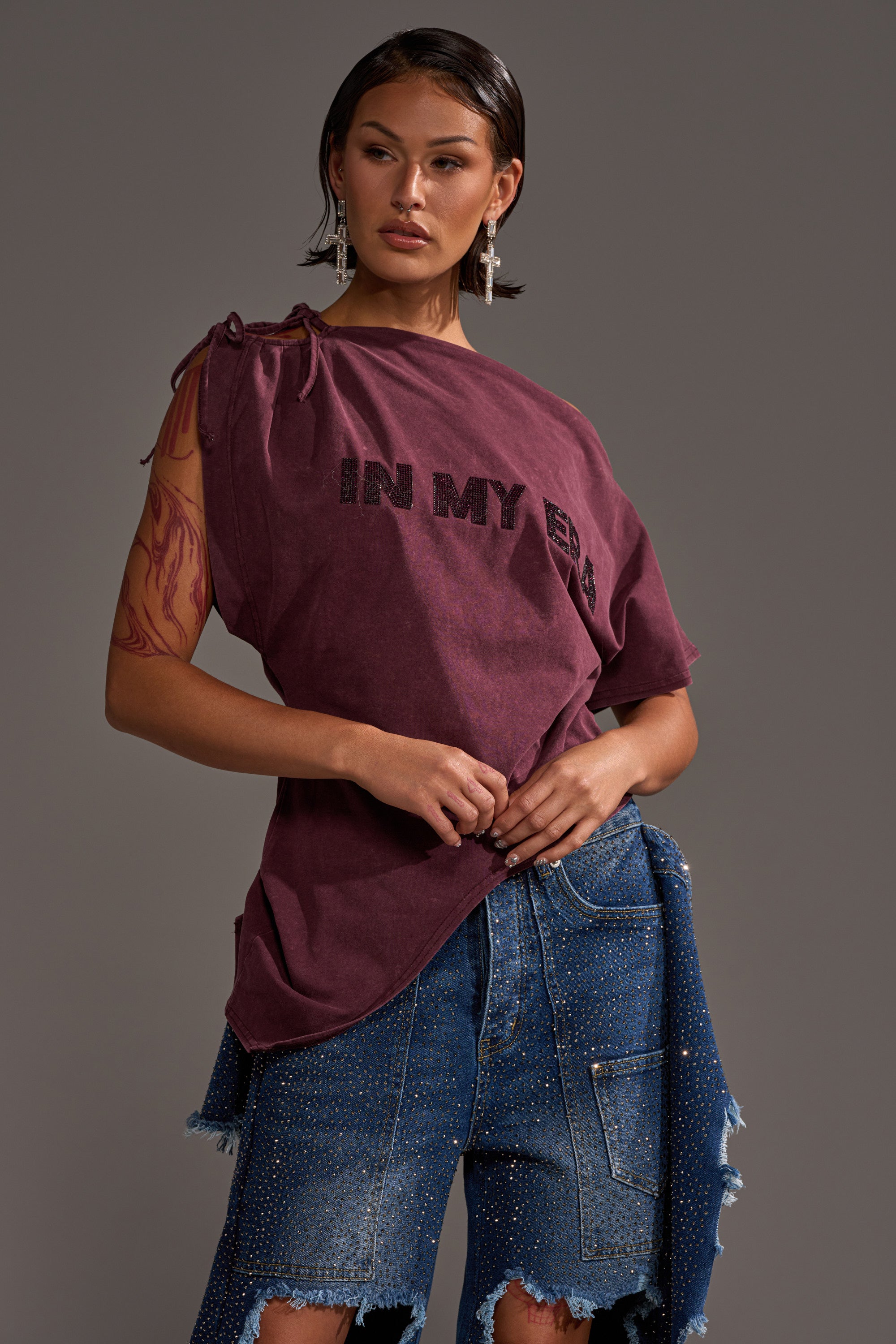 A woman poses confidently in the IN MY ERA OFF-THE-SHOULDER TEE paired with distressed denim shorts featuring rhinestone accents, accessorized with bold earrings, a temporary arm tattoo, and short wet-look hair.