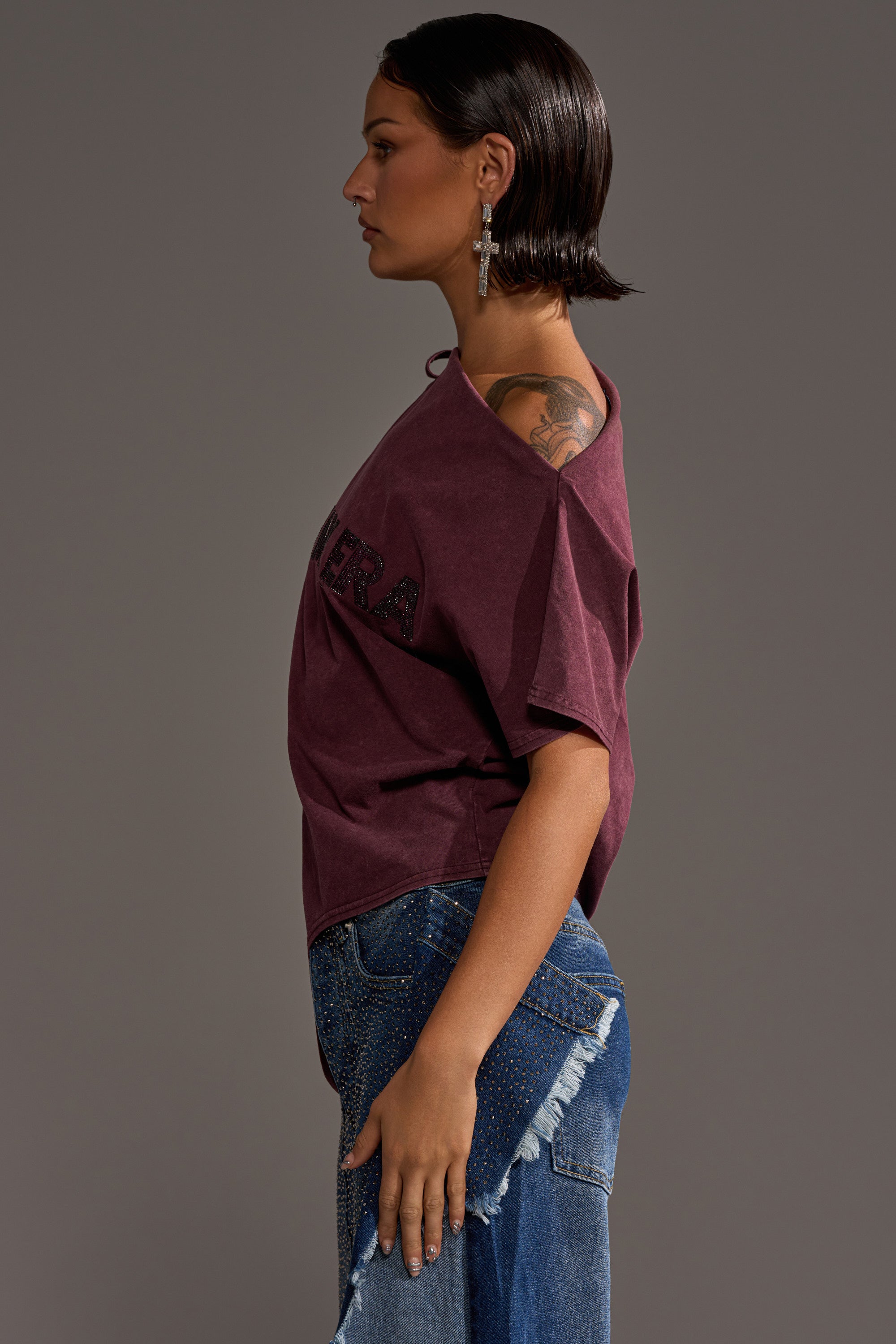 A woman with short dark hair in profile wears the IN MY ERA OFF-THE-SHOULDER TEE in maroon, paired with blue distressed jeans, large cross earrings, and reveals a shoulder tattoo against a plain background.