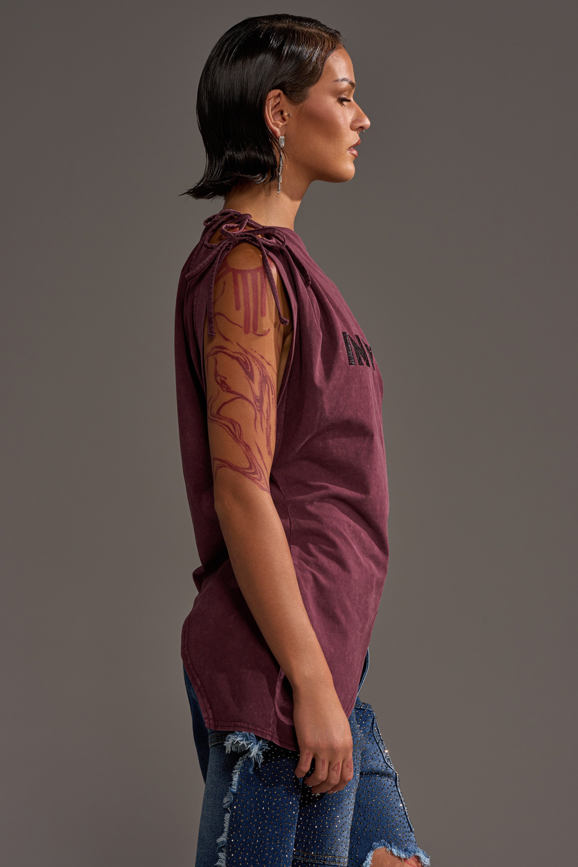 A woman with short dark hair and a large abstract tattoo on her left arm stands in profile against a gray background, wearing the IN MY ERA OFF-THE-SHOULDER TEE in maroon and ripped blue jeans.