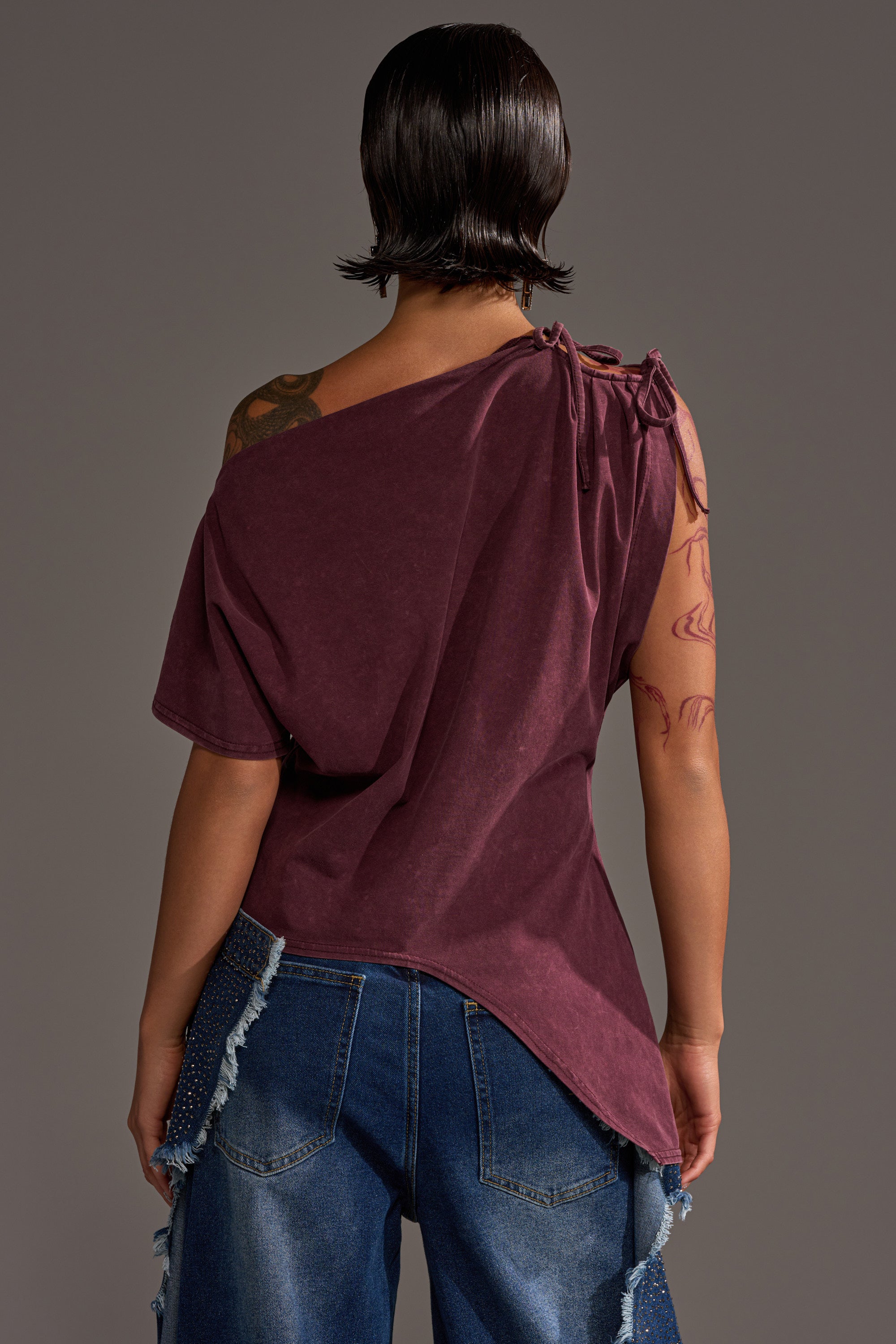 A person with short dark hair and visible arm tattoos is seen from the back in a burgundy IN MY ERA OFF-THE-SHOULDER TEE, paired with blue distressed jeans. The background is plain gray.