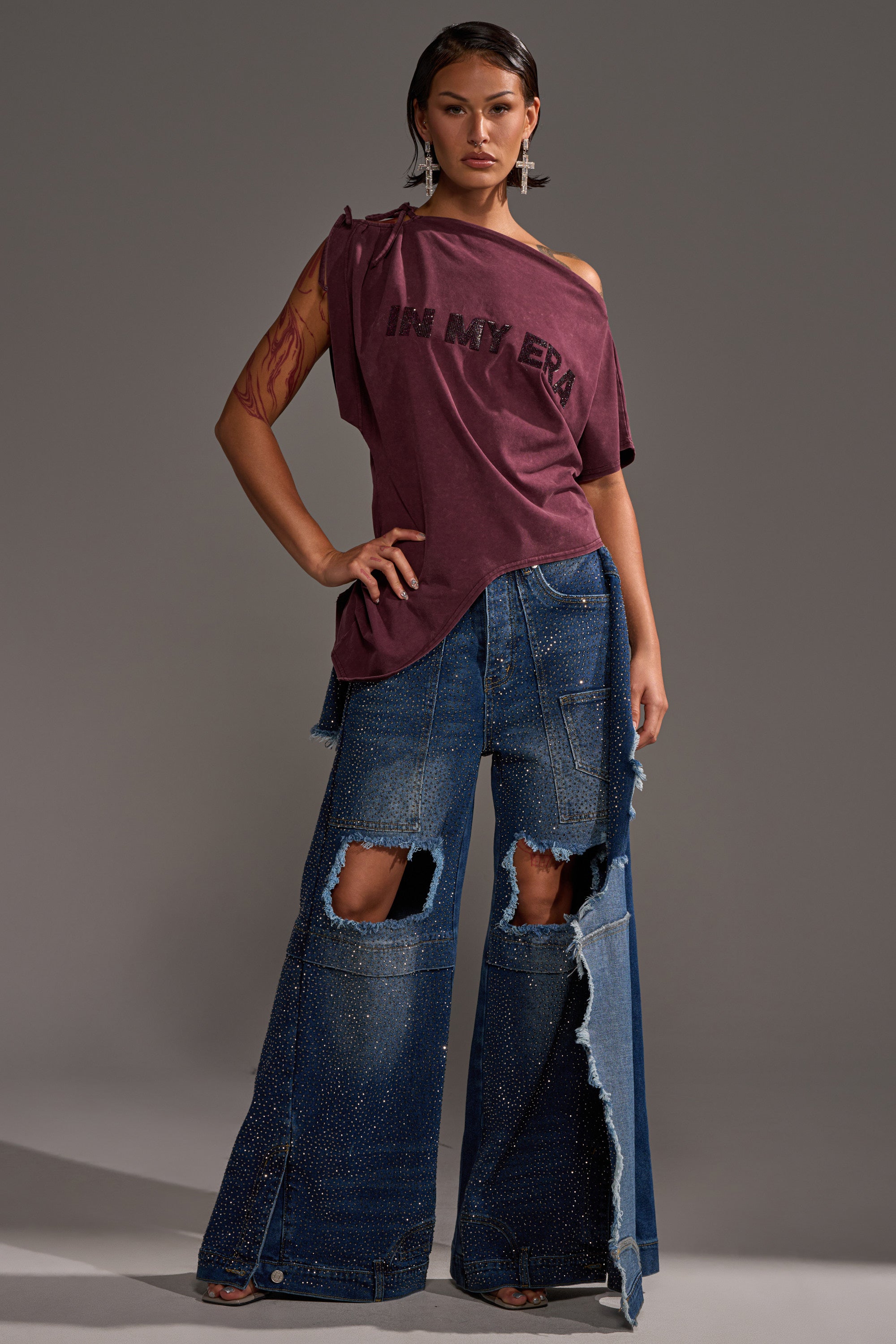A person stands confidently in the IN MY ERA OFF-THE-SHOULDER TEE and wide-leg, ripped, embellished blue jeans, accessorized with tattoos, earrings, and slicked-back hair against a gray backdrop.