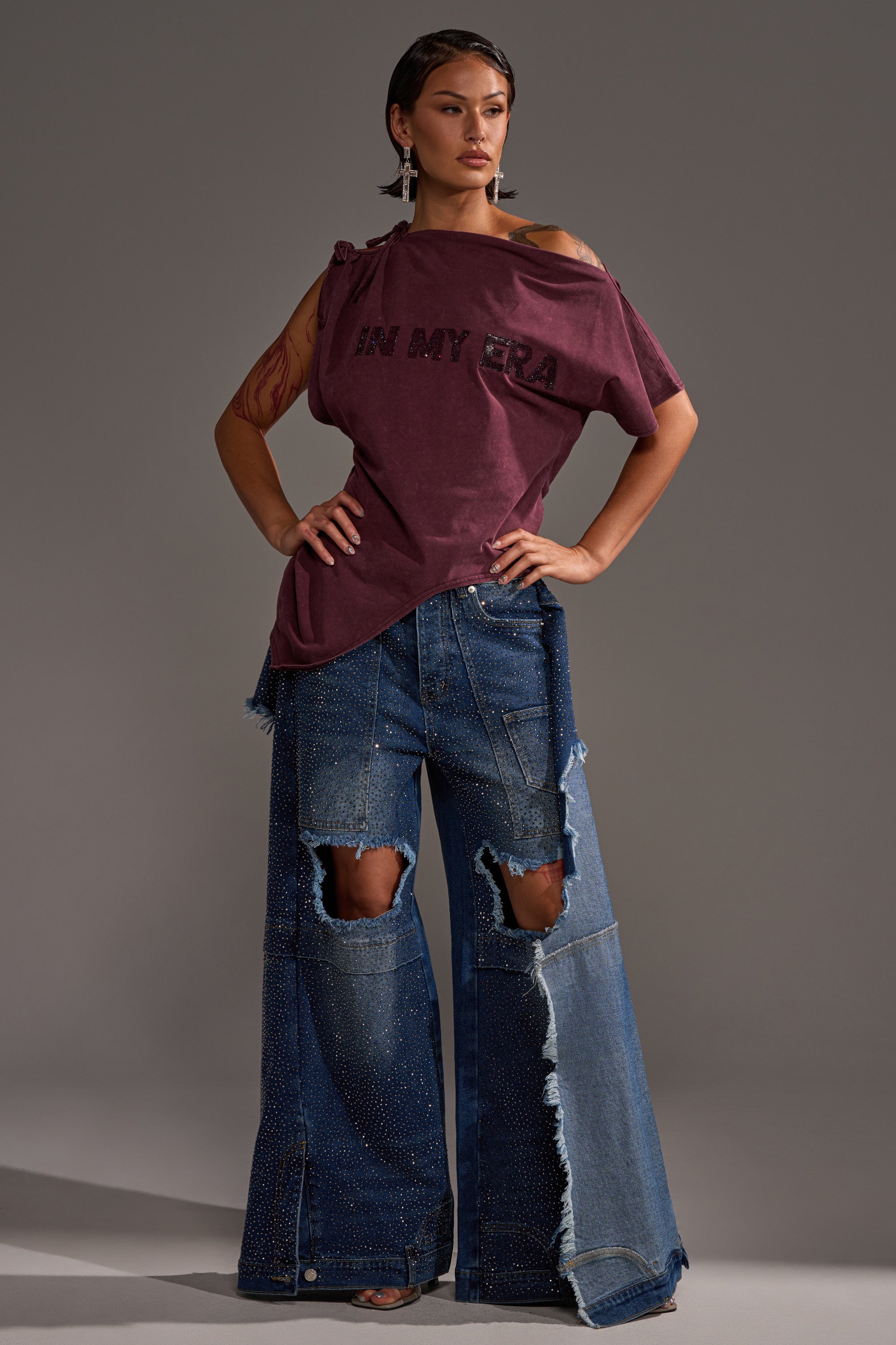 A woman poses confidently in the IN MY ERA OFF-THE-SHOULDER TEE in burgundy and wide-leg patchwork blue jeans with large knee rips and frayed hems, standing against a plain gray background.