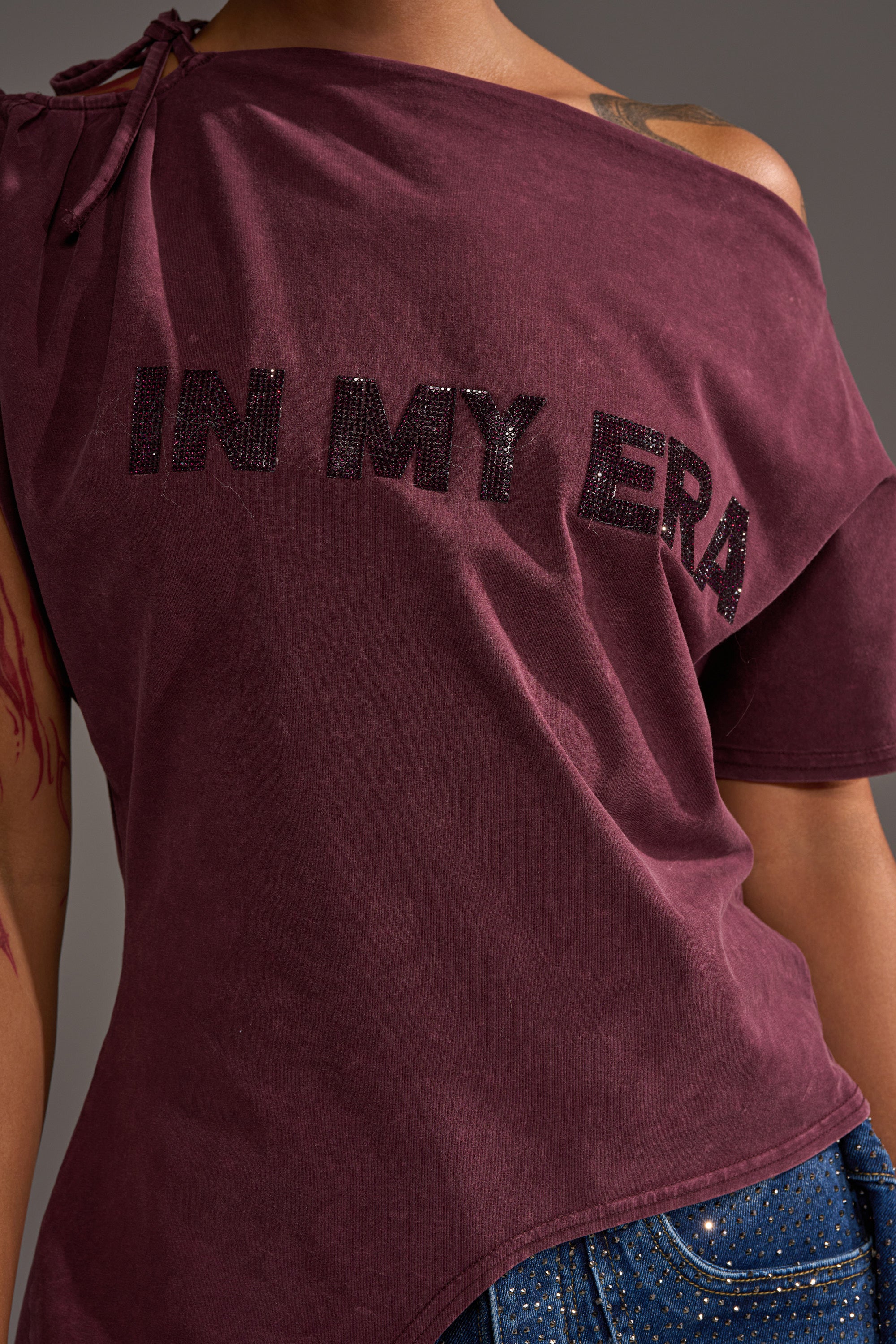 A person wears the IN MY ERA OFF-THE-SHOULDER TEE in maroon, featuring black beaded "IN MY ERA" lettering, paired with blue studded jeans. Only the upper body is visible.