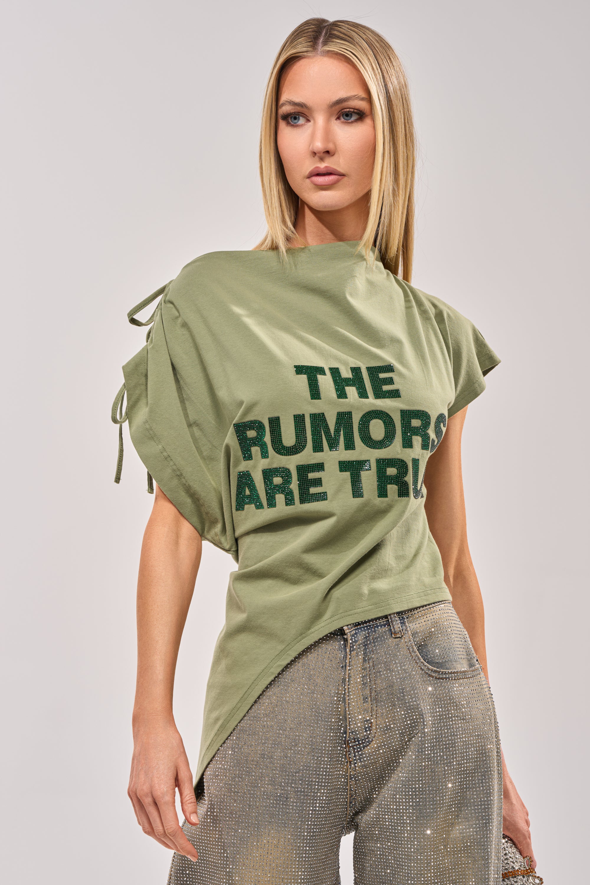A woman with straight blonde hair wears THE RUMORS ARE TRUE OFF THE SHOULDER OLIVE TEE and light blue rhinestone jeans, standing against a plain background. 0