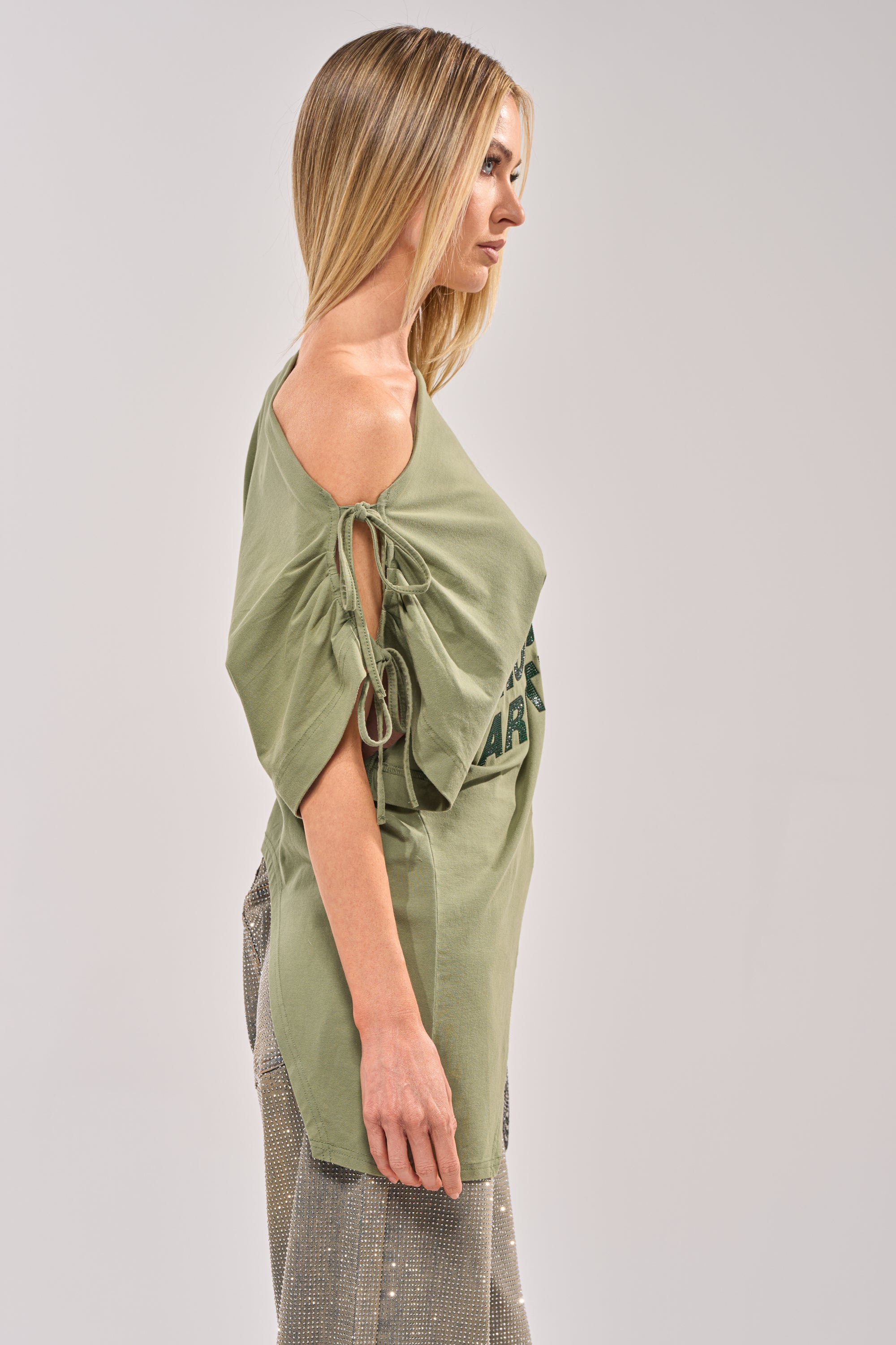 A woman with straight blonde hair poses in profile against a light gray background, wearing THE RUMORS ARE TRUE OFF THE SHOULDER OLIVE TEE with tie-sleeve details and loose shimmering pants for a chic streetwear look. 2