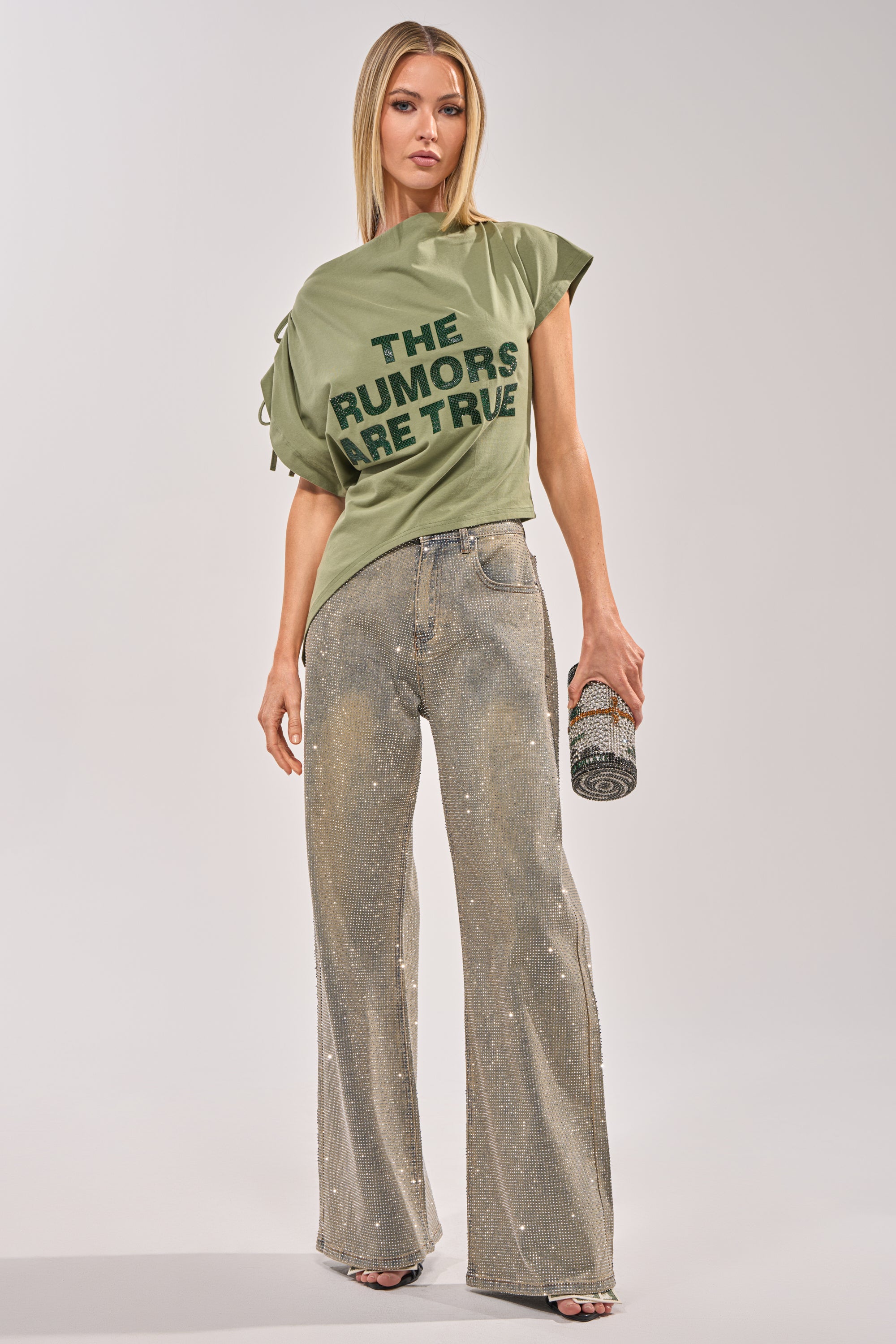 A woman faces forward in an off-the-shoulder olive tee with "THE RUMORS ARE TRUE" in bold black print, sparkling wide-leg jeans, and a glittery clutch—an eye-catching streetwear look. 5